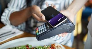 A person using a smartphone to make a contactless payment at a restaurant, with a plate of food in the foreground and a payment terminal seen on the table.